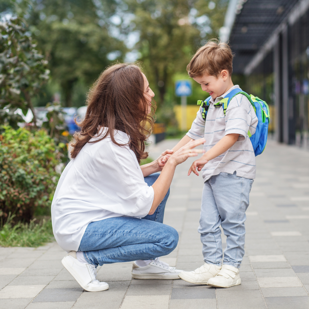 Man sieht eine braunhaarige Frau, die vor einem kleinen Jungen kniet und eine seiner Hände umfasst hält. Der Junge trägt einen kleinen Rucksack und lächelt.